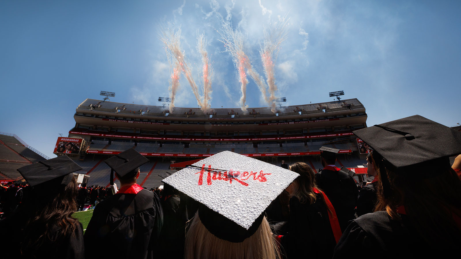 stadium on graduation day with fireworks and a student's cap that says Huskers on it